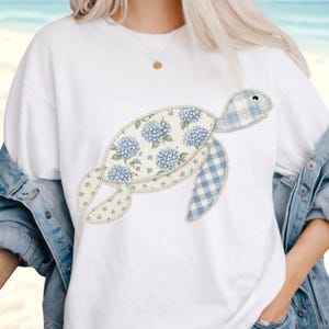 May include: White t-shirt featuring a whimsical turtle design. The turtle's body is adorned with blue hydrangea floral patterns and a blue gingham pattern. The design is stitched onto the shirt.