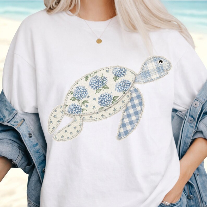 May include: White t-shirt featuring a whimsical turtle design. The turtle's body is adorned with blue hydrangea floral patterns and a blue gingham pattern. The design is stitched onto the shirt.