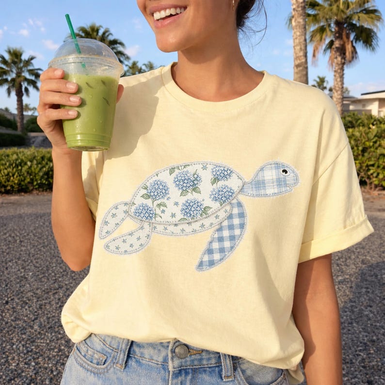 May include: A light yellow t-shirt featuring a turtle design. The turtle has a floral pattern on its shell and a blue and white checkered pattern on its flippers. The shirt is made of a soft material.