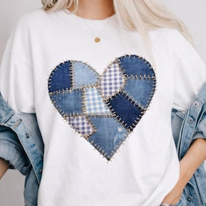 May include: White t-shirt featuring a large heart design made of denim patches in various shades of blue. The heart is stitched together with visible thread, creating a patchwork effect. The shirt is casual and comfortable.