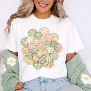 May include: White t-shirt featuring a large, colorful flower design. The flower is composed of various fabric circles in pastel shades of pink, green, and yellow, with floral and gingham patterns. The shirt is a casual, everyday wear item.