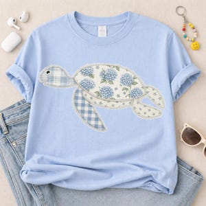 May include: A light blue t-shirt featuring a patchwork turtle design. The turtle's shell is adorned with blue hydrangea flowers and the flippers and head are patterned with a blue gingham check. The shirt has rolled-up sleeves.