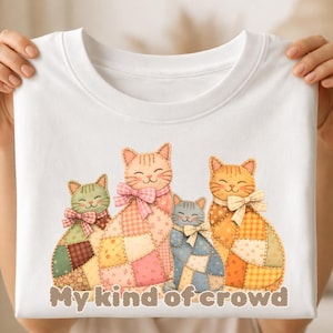 Könnte beinhalten: Weißes T-Shirt mit einer Grafik von vier lächelnden Patchwork-Katzen mit Schleifen. Die Katzen sind in den Farbtönen Orange, Rosa, Grün und Blau gehalten. Der Text "My kind of crowd" ist darunter in Braun gedruckt.