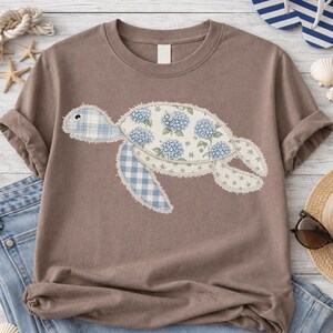 May include: A brown t-shirt featuring a patchwork turtle design. The turtle's shell is decorated with blue hydrangea flowers and the flippers and head are made of blue gingham fabric. The shirt has short sleeves and a crew neck.