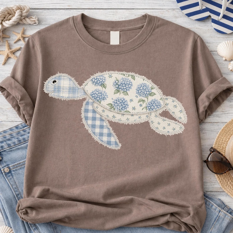 May include: A brown t-shirt featuring a patchwork turtle design. The turtle's shell is decorated with blue hydrangea flowers and the flippers and head are made of blue gingham fabric. The shirt has short sleeves and a crew neck.