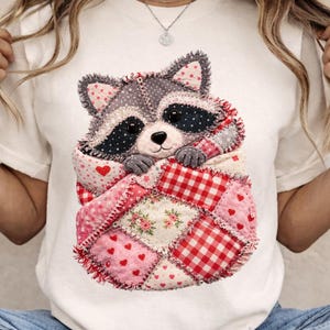 Peut inclure: T-shirt blanc avec un motif de raton laveur. Le raton laveur est niché dans une courtepointe patchwork avec des cœurs rouges, des motifs floraux et du vichy. Le raton laveur a un visage gris et noir avec des yeux noirs.