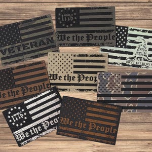 May include: A collection of rectangular patches featuring American flag designs in various colors and patterns. Some patches include text such as "VETERAN", "We the People", "1776", and "DON'T TREAD ON ME". The patches are displayed on a wooden surface.