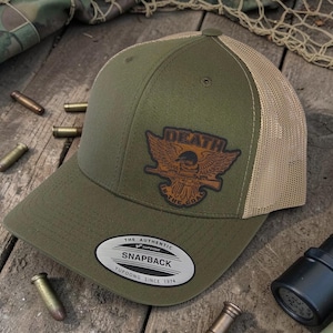 May include: An olive green snapback trucker hat with a tan mesh back. The front features a brown leather-like patch with an eagle graphic holding a rifle and the text DEATH IS THE GOAL. A circular sticker on the brim reads THE AUTHENTIC SNAPBACK.