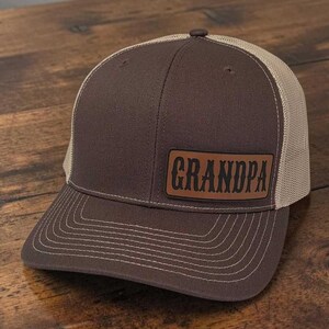 May include: A brown baseball cap with a tan mesh back and a brown leather-like patch on the front. The patch features the word GRANDPA in a dark, stylized font. The cap has a structured front panel and a curved brim with visible stitching detail.