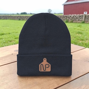 May include: A solid black knit beanie featuring a folded cuff. Centered on the cuff is a tan, tag-shaped leather patch embossed with the letters NP in a dark brown, sans-serif font. The beanie has a smooth, uniform texture throughout.
