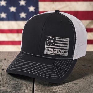 May include: A black and white trucker hat featuring a rectangular patch on the front. The patch displays a stylized flag design with a circle of stars, the number 250 in the center, and the text We the People printed in a bold, dark font.
