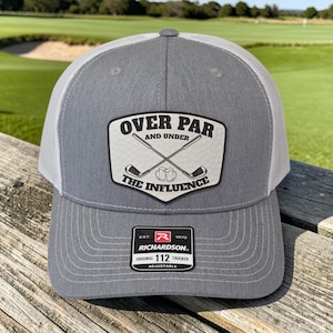 May include: A grey and white trucker hat featuring a patch with the text OVER PAR AND UNDER THE INFLUENCE, an illustration of two crossed golf clubs, and two beer mugs. A black Richardson 112 adjustable sticker is on the brim.
