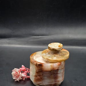 May include: A cylindrical stone container with a matching lid, featuring banded patterns in shades of brown, cream, and translucent white. The container is filled with a light pink granular substance and is accompanied by a small pink artificial flower.