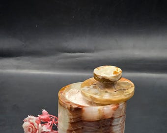 May include: A cylindrical stone container with a matching lid, featuring banded patterns in shades of brown, cream, and translucent white. The container is filled with a light pink granular substance and is accompanied by a small pink artificial flower.
