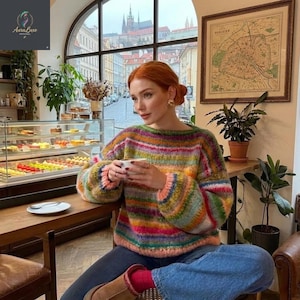 May include: A multi-coloured striped sweater in shades of pink, green, yellow, and blue. The sweater has a round neckline and long sleeves. The person is seated at a table in a cafe, holding a cup. The background features a window with a city view and a map.