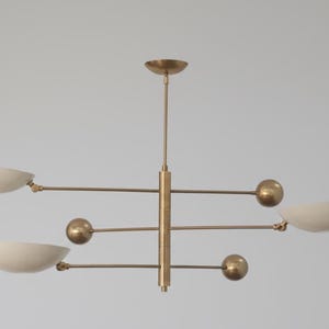 Puede incluir: Una lámpara de araña moderna de latón con cuatro pantallas en forma de cuenco de color blanco roto. La lámpara tiene una columna vertical central con brazos horizontales, cada uno con una pantalla y un adorno esférico. El diseño es minimalista y elegante.