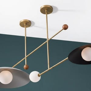 Puede incluir: Lámpara colgante moderna con acabado en latón, con tres pantallas ovaladas en negro y gris, cada una con una luz esférica. El diseño incluye detalles de madera y una estética minimalista, para interiores contemporáneos.