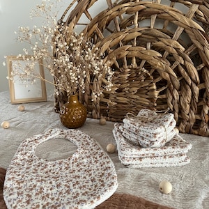 Puede incluir: Un babero para bebé y varias toallitas con un estampado floral en marrón y azul sobre fondo blanco. Una cesta de mimbre y un pequeño jarrón con flores secas también están en la imagen. Hay cuentas de madera esparcidas.