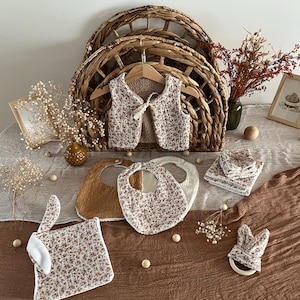 Puede incluir: Conjunto de artículos para bebé con estampado floral. Incluye un chaleco, baberos, toallitas y un mordedor con forma de conejo. Presentado sobre una superficie marrón con un fondo de cesta de mimbre.