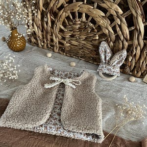 Puede incluir: Un chaleco de bebé beige y esponjoso con forro estampado floral y un lazo de encaje blanco. Una diadema a juego con orejas de conejo y una cesta de mimbre están en el fondo. La escena está sobre una tela de tonos neutros.