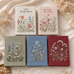 May include: Five rectangular fabric-covered journals featuring embroidered wildflower designs. Two journals have light-coloured covers with text: one reads 'Collect Beautiful Moments' and the other 'Live Simply Love Deeply'. Three journals have solid green, blue, and red covers.