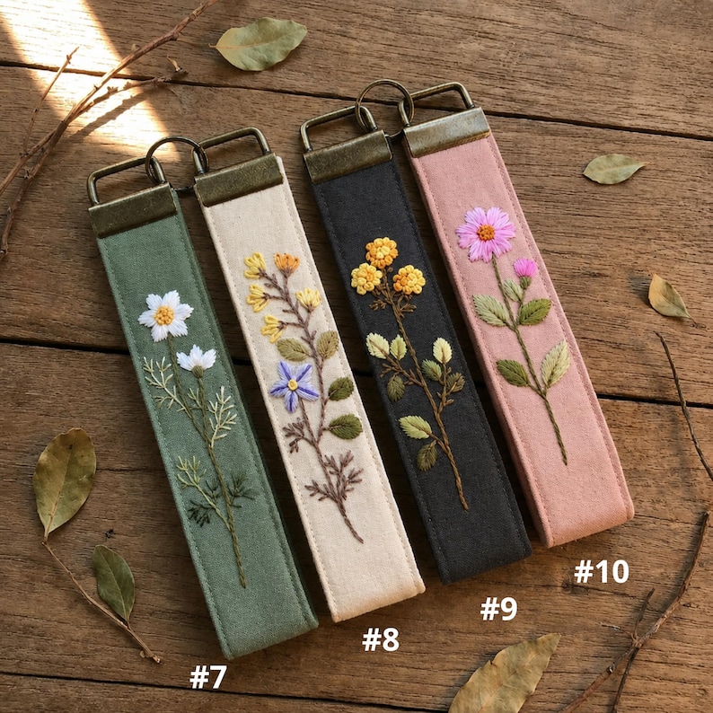 May include: Four rectangular fabric key fobs with bronze-toned metal hardware. From left to right: a sage green fob with white flowers, a cream fob with yellow and purple flowers, a charcoal gray fob with yellow flowers, and a dusty pink fob with a pink flower.