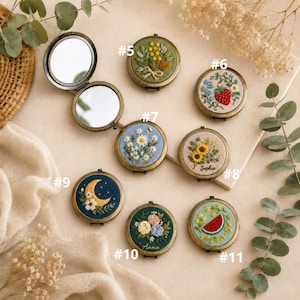 Op de afbeelding: Een collectie ronde metalen zakspiegels met geborduurde stoffen hoezen met diverse ontwerpen, waaronder bloemen, een aardbei, een maansikkel en een watermeloen, genummerd van 5 tot en met 11.