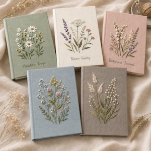 Könnte beinhalten: Fünf Notizbücher mit festem Einband aus Stoff in Salbeigrün, Creme, Altrosa, Hellblau und Grau. Jedes Notizbuch ist auf der Vorderseite mit gestickten Wildblumenmotiven verziert. Das grüne Buch trägt die Aufschrift Meadow Days, das cremefarbene Bloom Gently und das rosa Buch Botanical Journal.