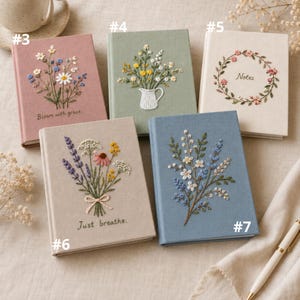 May include: Five rectangular fabric-covered journals featuring embroidered floral designs. The journals are coloured dusty pink, sage green, cream, beige, and slate blue. Text on the covers includes "Bloom with grace," "Notes," and "Just breathe."