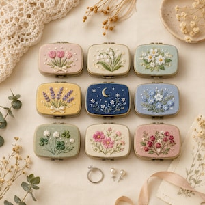 Puede incluir: Nueve estuches metálicos rectangulares con tapas de tela bordada que presentan diseños florales, botánicos y celestiales en varios colores incluyendo rosa, amarillo, azul, verde y crema, dispuestos en cuadrícula con un anillo y dos pendientes.