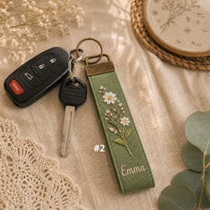 May include: A rectangular olive green fabric keychain featuring an embroidered floral design of white daisies and small pink buds, with the name Emma stitched in white thread at the bottom, attached to a bronze-toned metal ring with two car keys.