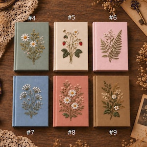May include: Six rectangular notebooks with embroidered botanical covers. #4 is sage green with daisies, #5 is cream with strawberries, #6 is pink with ferns, #7 is blue with white flowers, #8 is coral with daisies, and #9 is tan with wildflowers.