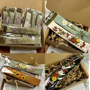 May include: A collection of fabric key fobs featuring intricate floral and botanical embroidery. Each rectangular strap is finished with a bronze-toned metal clasp and ring. The designs include lavender, daisies, sunflowers, and mushrooms.