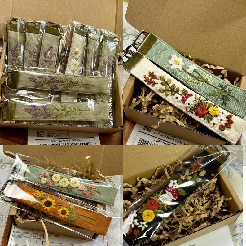 May include: A collection of fabric key fobs featuring intricate floral and botanical embroidery. Each rectangular strap is finished with a bronze-toned metal clasp and ring. The designs include lavender, daisies, sunflowers, and mushrooms.