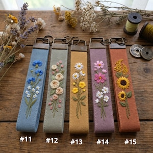 May include: Five rectangular fabric key fobs featuring floral embroidery. From left to right: blue with cornflowers, sage green with roses, mustard yellow with daisies, lavender with cosmos, and terracotta with sunflowers. Each has a metal ring.