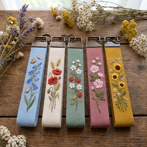 Könnte beinhalten: Fünf rechteckige Schlüsselanhänger aus Stoff mit bronzefarbenen Metallbeschlägen. Jeder zeigt eine einzigartige Stickerei: blaue Glockenblumen, rote Mohnblumen mit Weizen, weiße Erdbeerblüten mit roten Beeren, rosa Wildrosen und gelbe Sonnenblumen.