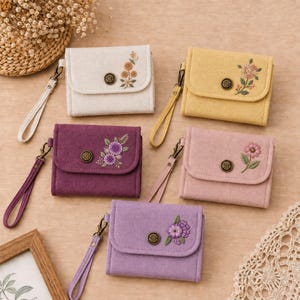 May include: Five rectangular fabric wallets in cream, yellow, deep purple, light pink, and lavender. Each wallet features a flap with a central metallic button, a matching wrist strap, and a unique floral embroidery design on the front flap.