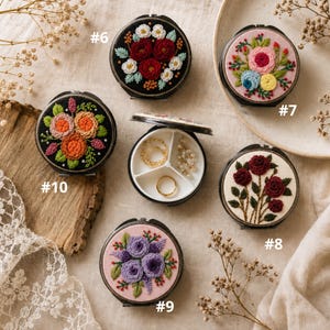 Op de afbeelding: Een collectie ronde, scharnierende metalen doosjes met geborduurde bloemmotieven op de deksels, genummerd van #6 tot #10. Eén doosje staat open en toont een witte binnenkant verdeeld in drie vakjes met kleine gouden ringen en kralen.