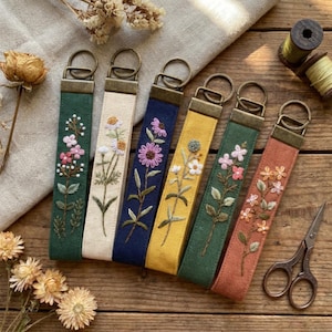 Könnte beinhalten: Sechs Schlüsselanhänger aus Stoff mit gestickten Blumenmotiven in verschiedenen Farben wie Grün, Creme, Marineblau, Gelb und Terrakotta. Jeder Anhänger hat eine bronzefarbene Metallhalterung mit einem Schlüsselring.