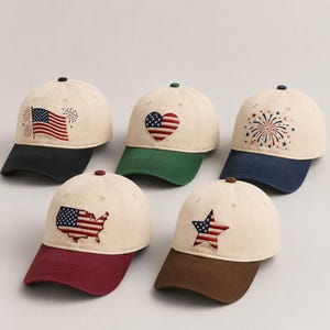 Puede incluir: Cinco gorras de béisbol con coronas de color crema y viseras en contraste. Las gorras presentan diseños bordados que incluyen una bandera estadounidense con fuegos artificiales, un corazón, una explosión de fuegos artificiales, un mapa de los Estados Unidos y una estrella, todo en rojo, blanco y azul.