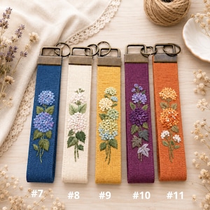 May include: Five rectangular fabric key fobs with bronze-toned metal hardware and split rings. Each features unique floral embroidery: blue, white, yellow-green, purple, and orange hydrangea designs. Labeled #7, #8, #9, #10, and #11.