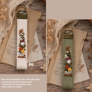 May include: Two fabric keychains with bronze-toned metal hardware. One is off-white and the other is sage green. Both feature vertical embroidery of a red-capped mushroom, orange and yellow flowers, and brown leaves. The text #6 is visible.