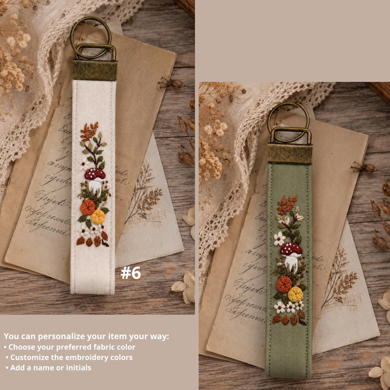 May include: Two fabric keychains with bronze-toned metal hardware. One is off-white and the other is sage green. Both feature vertical embroidery of a red-capped mushroom, orange and yellow flowers, and brown leaves. The text #6 is visible.