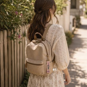 May include: A beige canvas backpack with a top handle and a front zippered pocket. A rectangular fabric keychain with a lavender floral print and the text "Sophia" hangs from the zipper pull. The backpack features brown accents.