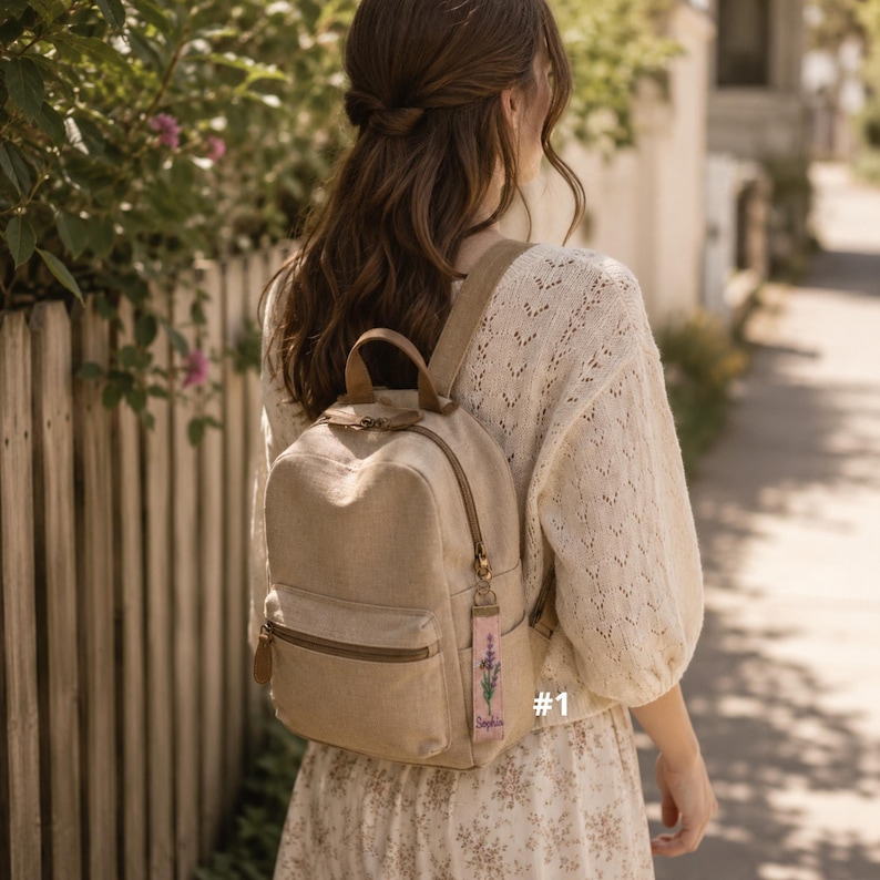 May include: A beige canvas backpack with a top handle and a front zippered pocket. A rectangular fabric keychain with a lavender floral print and the text "Sophia" hangs from the zipper pull. The backpack features brown accents.