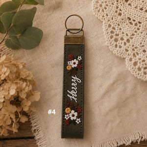 May include: A dark olive green fabric keychain featuring an embroidered floral pattern in white, yellow, and rust red, with the name "Herry" stitched in white cursive thread. The keychain includes a bronze-toned metal clasp and ring.