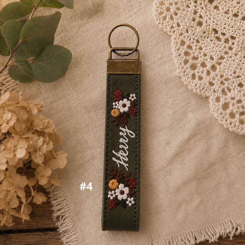 May include: A dark olive green fabric keychain featuring an embroidered floral pattern in white, yellow, and rust red, with the name "Herry" stitched in white cursive thread. The keychain includes a bronze-toned metal clasp and ring.