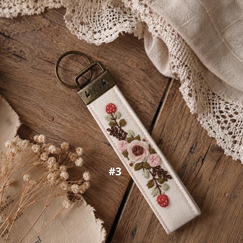 May include: A cream-colored fabric keychain featuring an embroidered floral design with pink roses, green leaves, and red berries. The keychain is attached to a bronze-toned metal hardware ring and clasp. The number 3 is printed on the fabric.