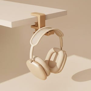 Può includere: Cuffie over-ear beige appese a un morsetto in legno fissato a una superficie bianca. Le cuffie hanno un design minimalista con grandi padiglioni e un archetto imbottito. Lo sfondo è di colore beige uniforme.