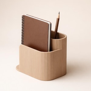 Op de afbeelding: Een beige bureau-organizer met een verticale geribbelde textuur en twee vakken. Het ene vak bevat een donkerbruin notitieboek met spiraalbinding en het andere vak bevat een enkel donkerbruin houten potlood dat rechtop in de houder staat.
